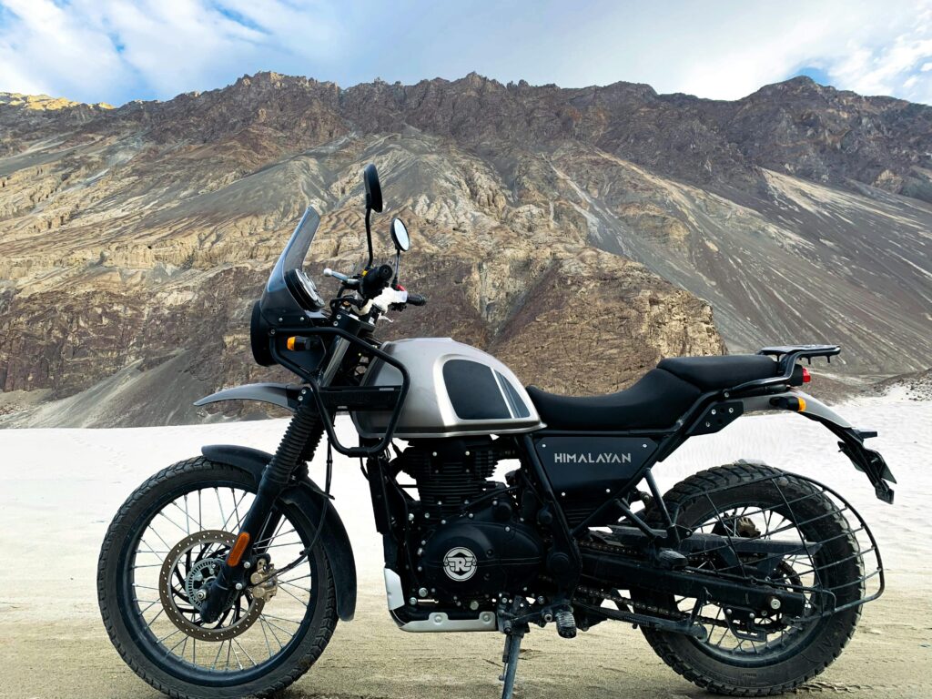 Royal Enfield Himalayan adventure motorcycle parked on a high-altitude mountain terrain with rocky peaks backdrop.