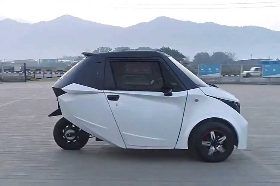 खुले पक्के क्षेत्र में सफेद रंग की Strom Motors R3 छोटी इलेक्ट्रिक कार साइड प्रोफाइल में खड़ी दिखाई दे रही है, जिसके पीछे पहाड़ और सड़क का दृश्य है।