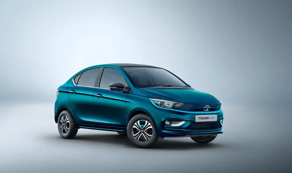 स्टूडियो जैसे हल्के ग्रे बैकग्राउंड में नीले रंग की Tata Tigor EV इलेक्ट्रिक सेडान सामने की ओर तिरछे एंगल में खड़ी दिखाई दे रही है।