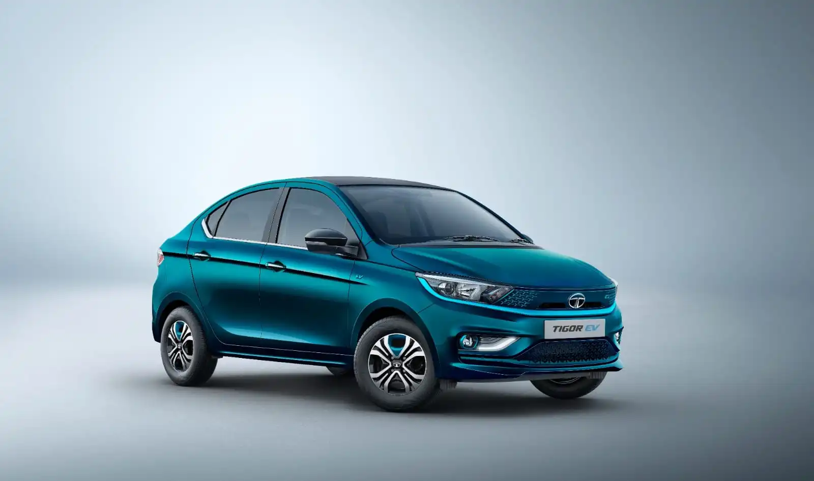 स्टूडियो जैसे हल्के ग्रे बैकग्राउंड में नीले रंग की Tata Tigor EV इलेक्ट्रिक सेडान सामने की ओर तिरछे एंगल में खड़ी दिखाई दे रही है।